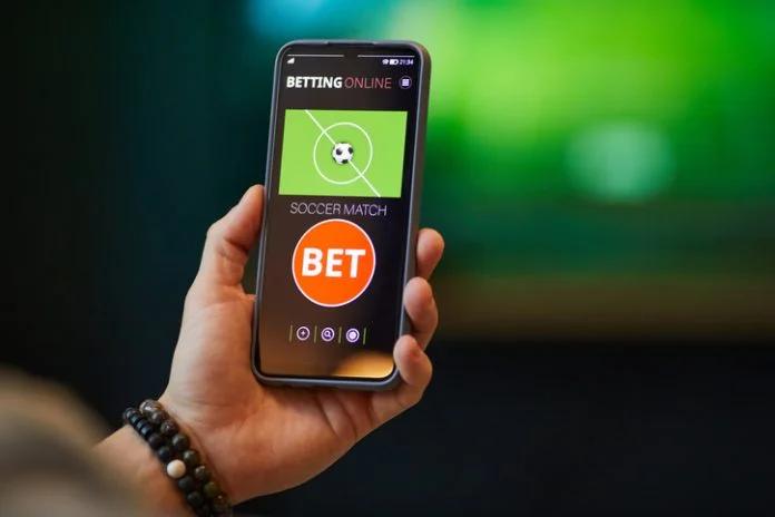มือกำลังถือสมาร์ทโฟนที่เปิดหน้าแอปพนันบอลออนไลน์ พร้อมปุ่ม BET บนหน้าจอ ใช้ประกอบสอน วิธีแทงบอลผ่านมือถือ แบบง่ายสำหรับผู้เริ่มต้น